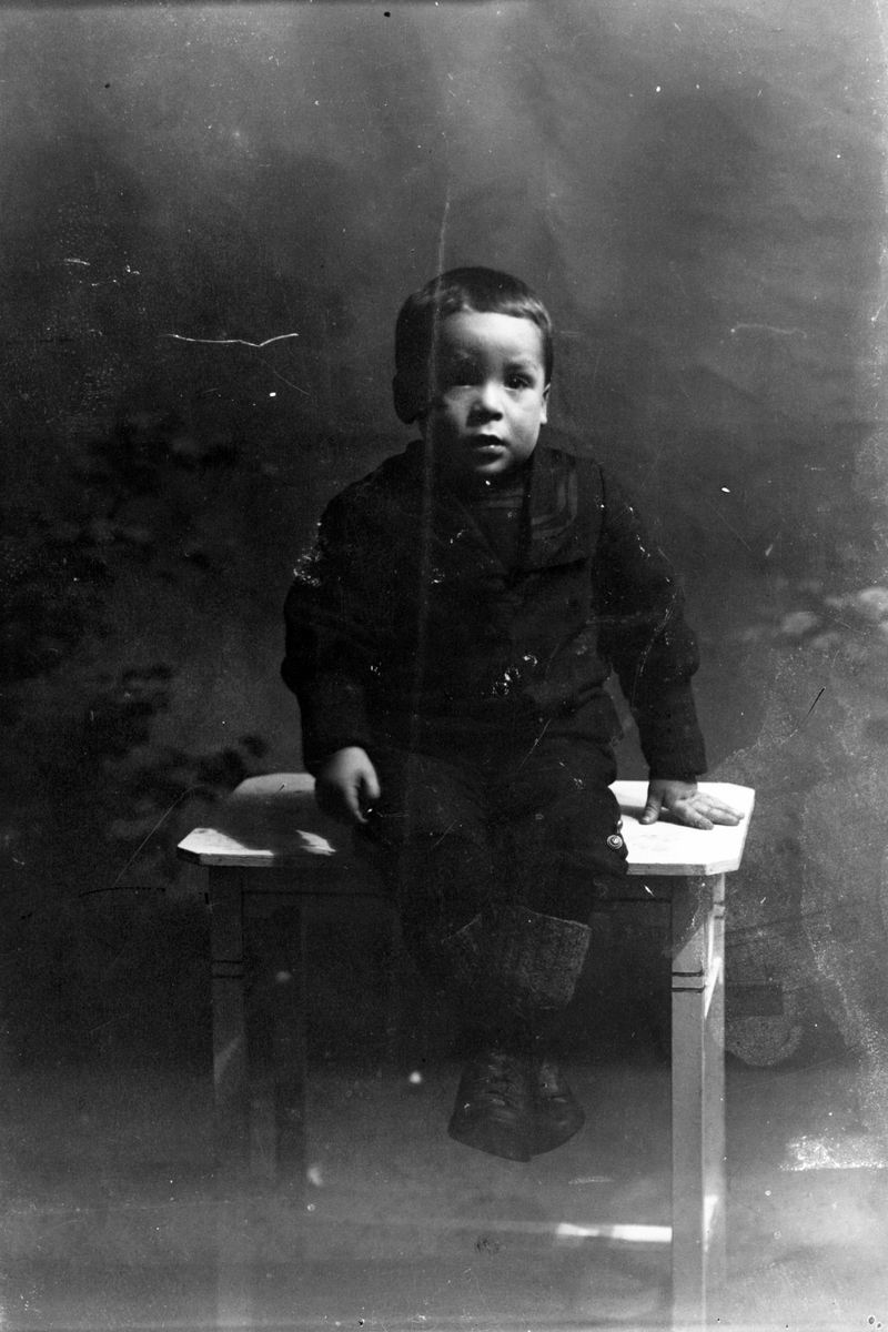 Studioportrett av en liten gutt sittende på et bord.
