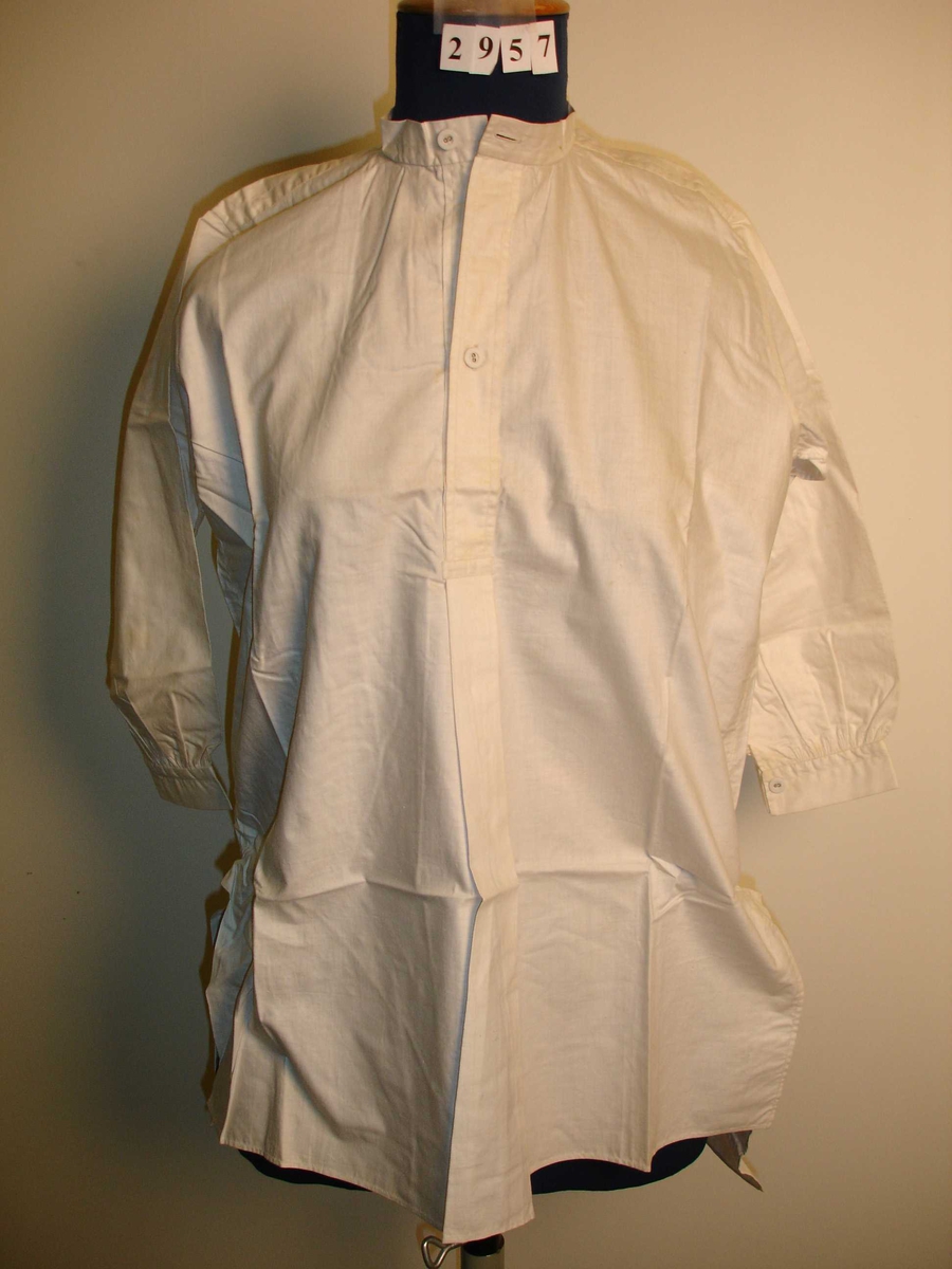 Skjorte, langermet, kortere foran enn bak, skjorten har mange små rynker bak i nakken og ved mansjettene på ermene. Kort stolpe foran med knepping.  Hvitt bomullstoff. 
