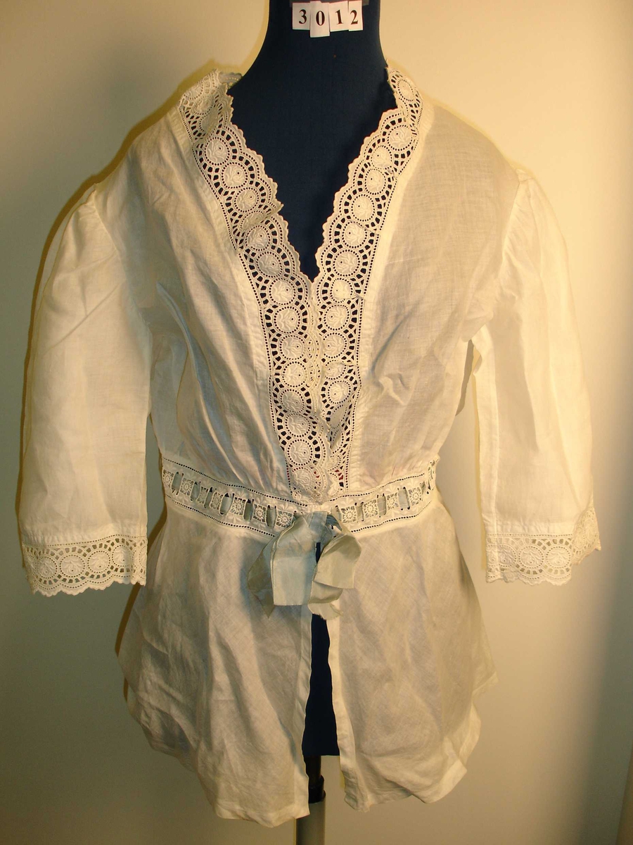 Frisørjakke, bluse, skjorte i hvit bomullstoff, blonderkant rundt ermene, langs hals, midje og front, I midjen er det tredd inn blå silkebånd. Lukkes med trykknapp