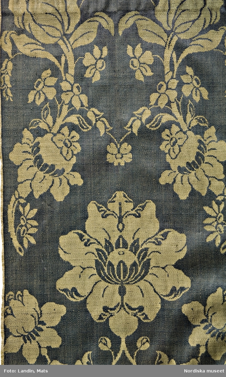Förkläde av stormönstrad ylledamast i 1700-talsmönstring. Gulgrönt stort blommönster på mörkgrön botten. Förklädet är hopsytt av 2 hela vådbredder med stadkanter i var sida. Upptill rynkat mot en remsa av mörkblå vadmal med fastsydda knytband av olika hemvävda band. I ena sidan ett ripsband i brunt, blått och gult ullgarn på linvarp och i den andra sidan ett mönstervävt band med rött upplockat mönster på oblekt linnebotten. I mönstret är invävt initialerna L K O D. I nederkanten är förklädet skott med 3,5 cm brett ylleband skarvat av ett helrött band och ett rött med smala vita ränder. På insidan av midjelinningen broderat med vitt: 90.
Förklädet är unikt och mycket välbevarat sånär som på några malhål på midjeremsan av vadmal.
Ylledamast av denna höga kvalitet vävdes i första hand i Norwich, England, men tillverkning fanns även på andra platser eftersom detta var ett modernt tyg under 1700-talet. Till Sverige kom utländska tyger av denna sort in i allmogedräkten i slutet av 1700-talet och eftersom det var dyrbara köpetyger användes de till högtidsdräkt i första hand bröllopsdräkterna för både kvinnor och män, vanligast som livstycken och västar, i enstaka fall som kjolar och ännu mer sällan som förkläden.  I detta fall kan man se hur rapporten har sett ut, vilket är svårare på de tillskurna livstyckena. Eftersom sådana utländska tyger hade importförbud under 1700-talet kan de ha kommit illegalt via gränshandeln med Norge.
Förklädet kom in tillsammans med ett par sammanbundna kjolväskor av  rött tyg med påsydda silverband, inv.nr  98867 som använts som brudklädsel. Det kan mycket väl vara från samma brud.
/Berit Eldvik 2008-06-09

I sin bok "Möte med mode" (2014), sid 120-121, skriver Berit Eldvik att förklädet är vävt i Norwich.