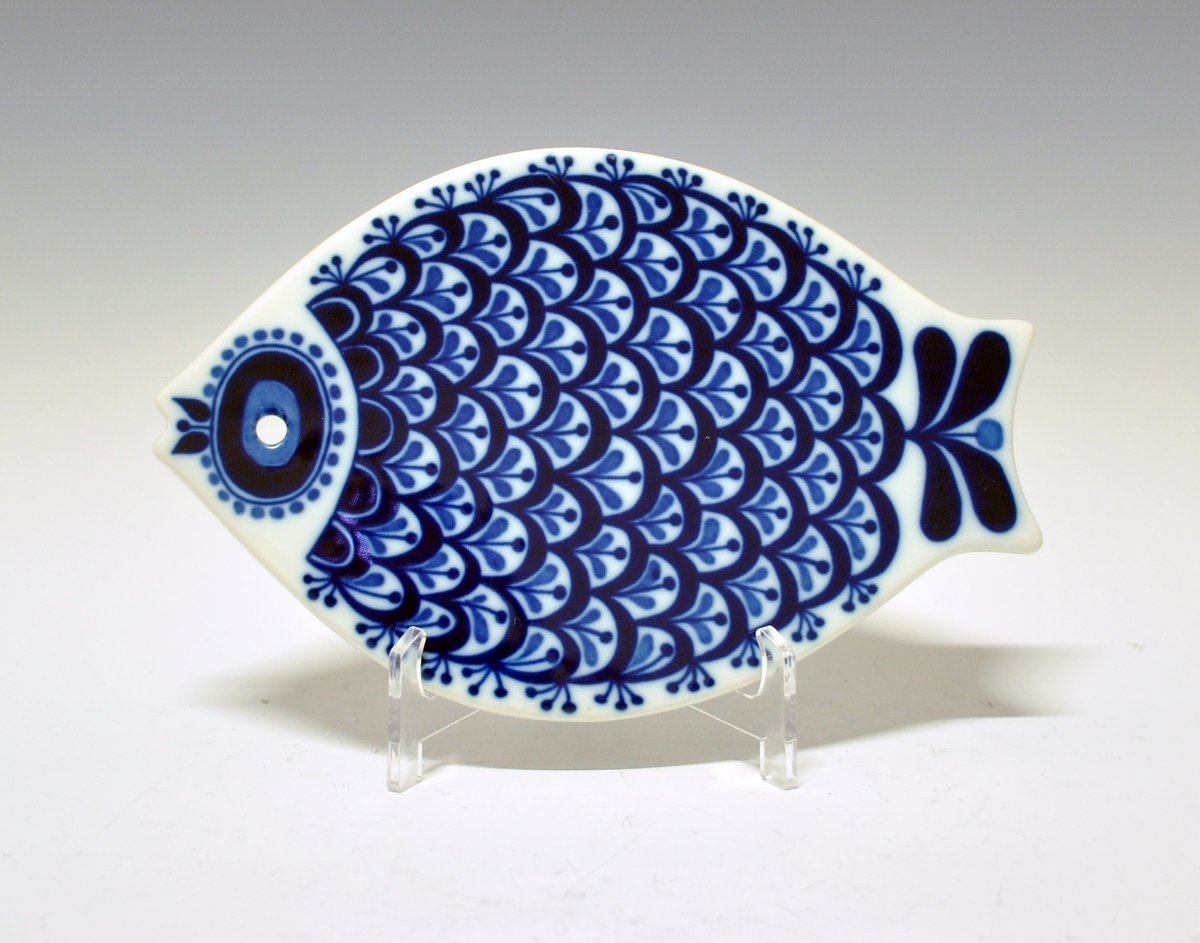 Smørefjel i porselen. Formet som fisk med hull til oppheng. Hvit glasur. Dekorert med stilisert plantemønster i blått. 
Modell: Arne Lindaas.
Dekor: Beth Breyen