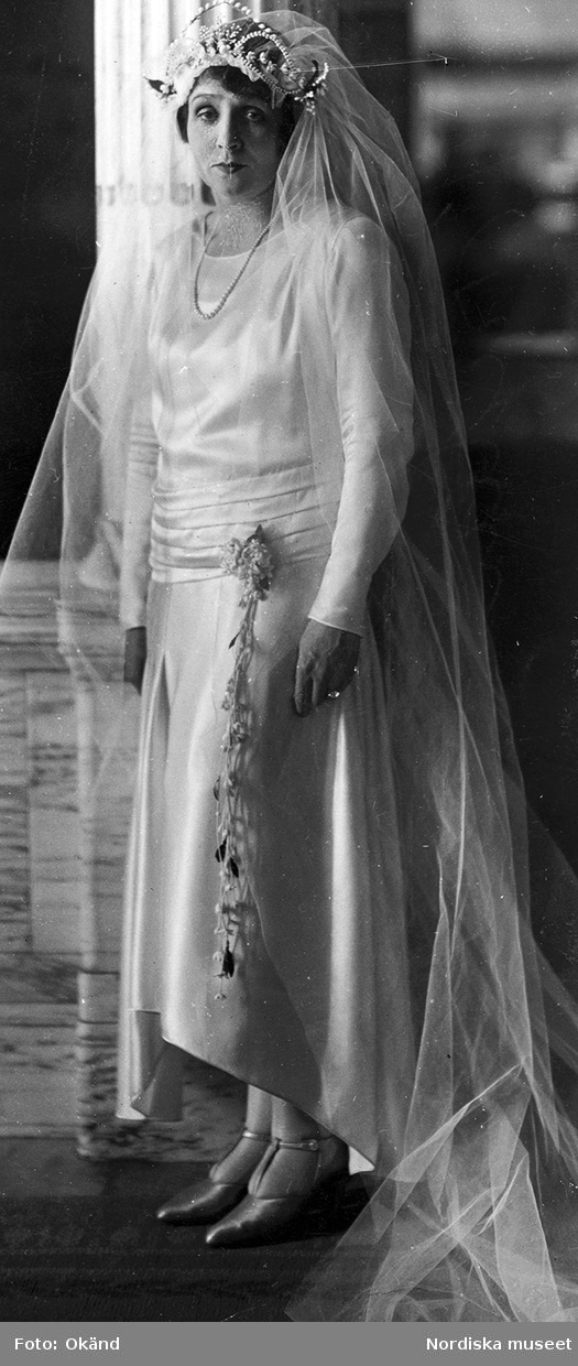 Fru Maria Hedberg i brudklänning med lång slöja, dekorerad med pärlor och garnityr, blomsterdekoration hängande från höften.