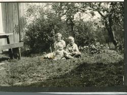 Tre barn sitter i gresset i hage. Benk og bord ved husveggen