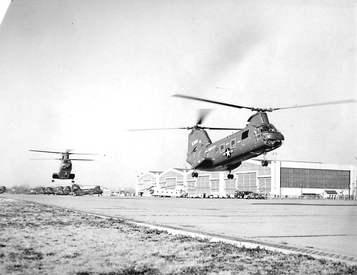 2 helikoptre i luften. Boeing Sea Knight UH-46D. Hangar og flere helikoptere i bakgrunnen.