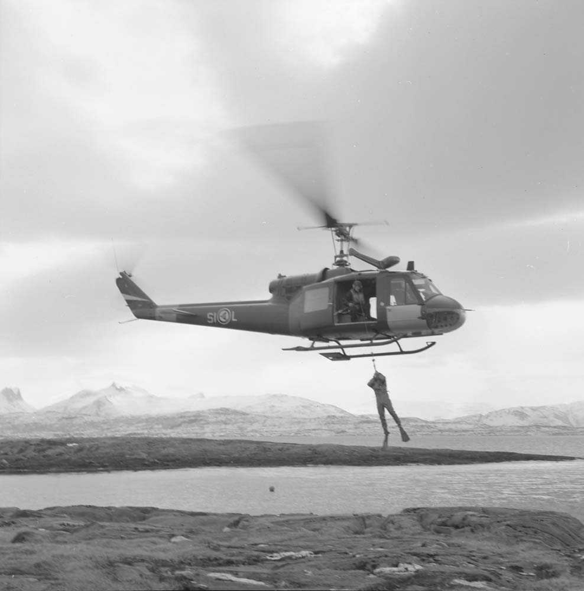 UH-1B (Bell 204), tilhørende 717 skvadron, med froskemann svevende over Bodø flystasjon, med Børvasstindan i bakgrunnen.