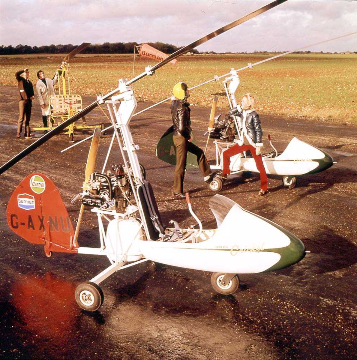 To helikopter på bakken, Campbell Cricket Autogyro. To personer står ved det ene helikoptret. To personer i bakgrunnen.