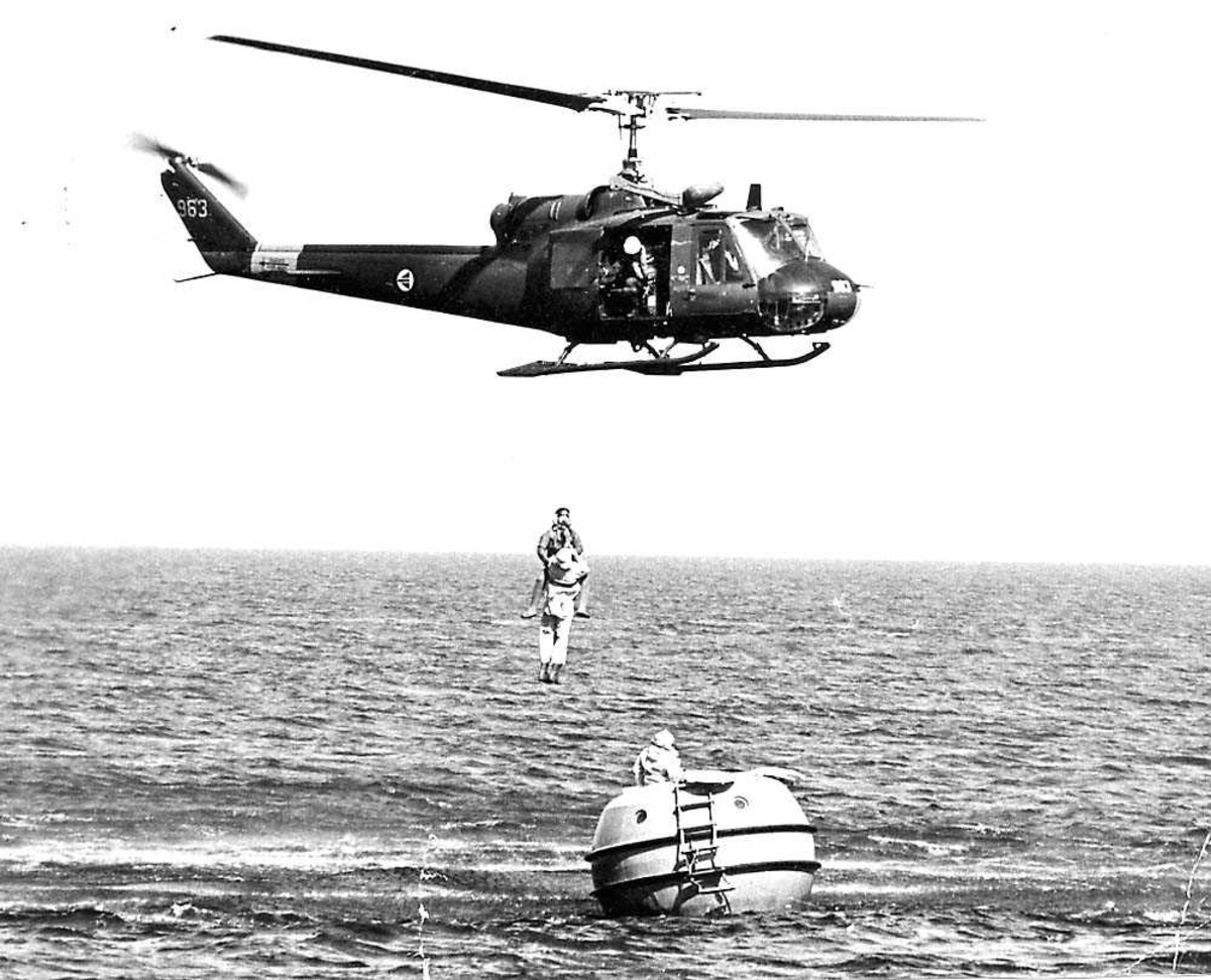 Luftfoto. Ett helikopter i luften, Bell UH-1B (204B) under en redningsøvelse. To personer henger under helikoptret. En redningsflåte på vannet.