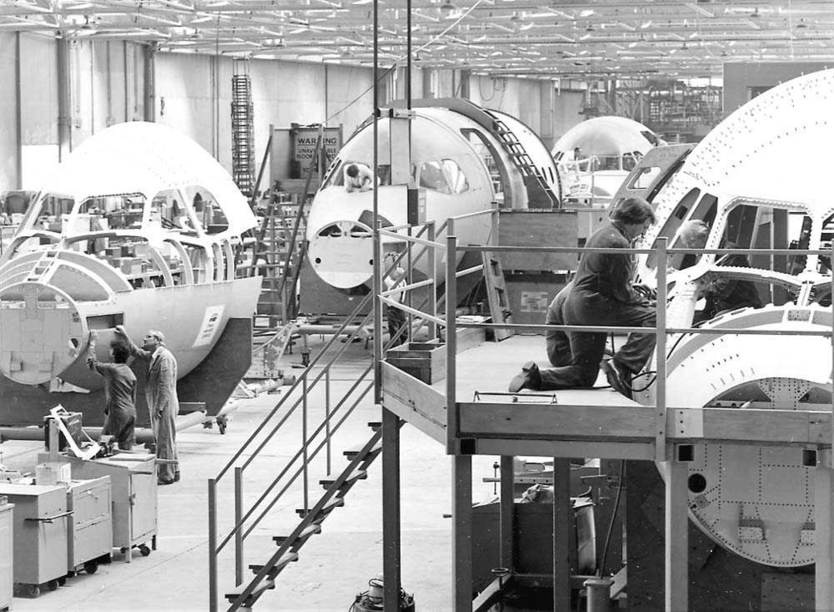 Flere nesepartier av BAe 146 inne i en flyfabrikk, som klargjøres til ferdigmontering. Flere personer som arbeider ved flyene.