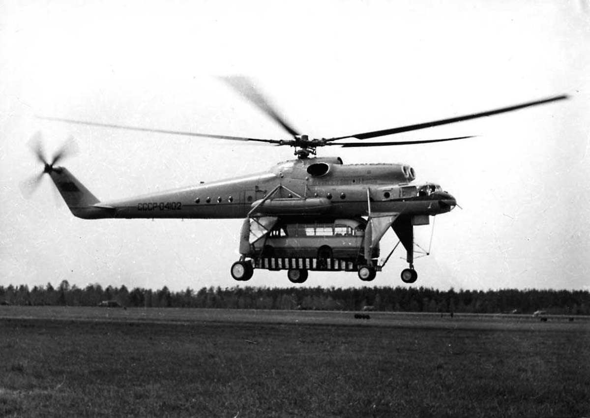 Ett helikopter i luften like over bakken, Mil Mi-10 (V-10) "Harke" flying crane. En buss henger under helikoptret.