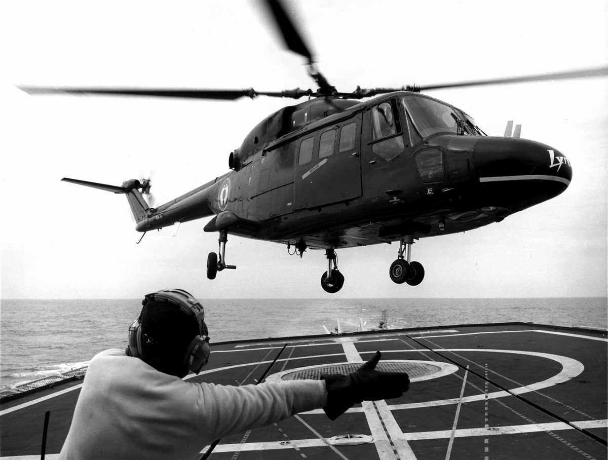 Ett helikopter i luften, like over helipad på skipsdekk, en person i forgrunn, dirigerer landing. Westland Lynx HAS Mk.2