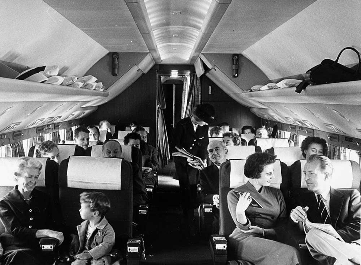 Interiøret inne i en flykabin. Flere passasjer som sitter i stolene, en flyvertinne i midtgangen.