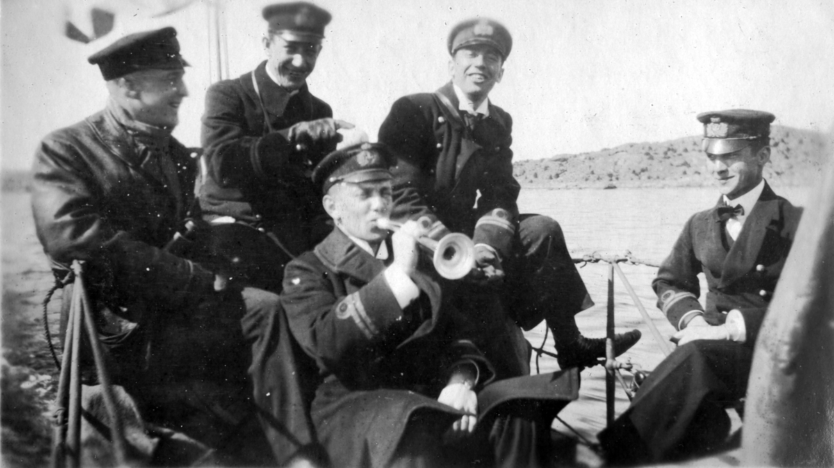Fem sjöbefäl sitter på fartygs däck där en av de spelar trumpet. Alla bär sjöbefälsuniformerer och hattar.