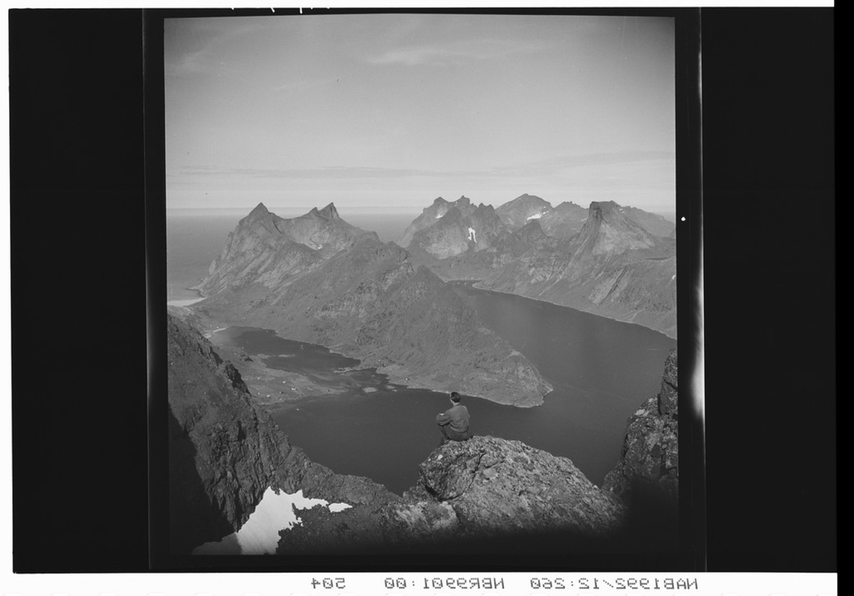 Mann sitter på bergknaus og speider utover fjellformasjon. Hav og fjord rundt.