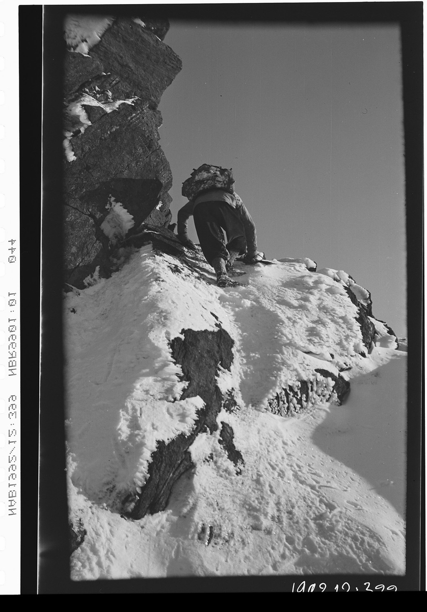 Mann med ryggsekk klatrer i snødekt fjellskråning.