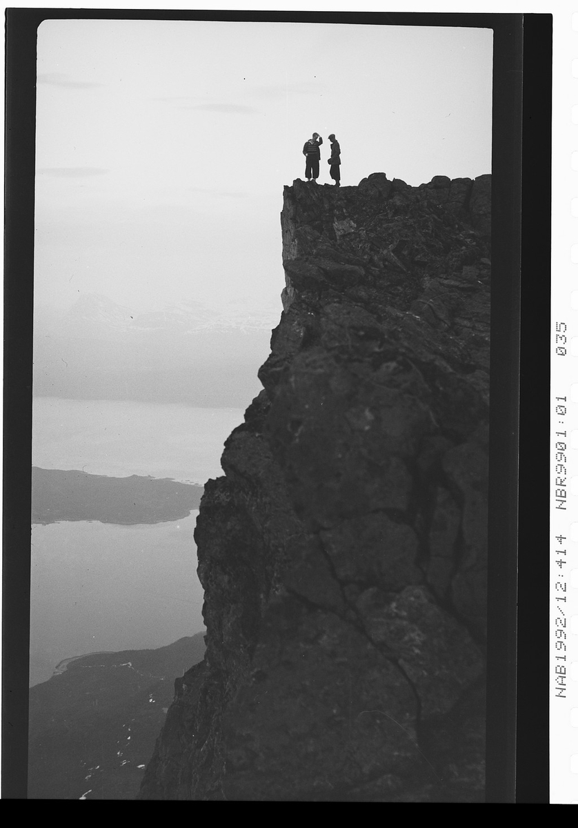 To menn står ved kanten av en høy klippe, dal og sjø i bakgrunnen. Ukjent.