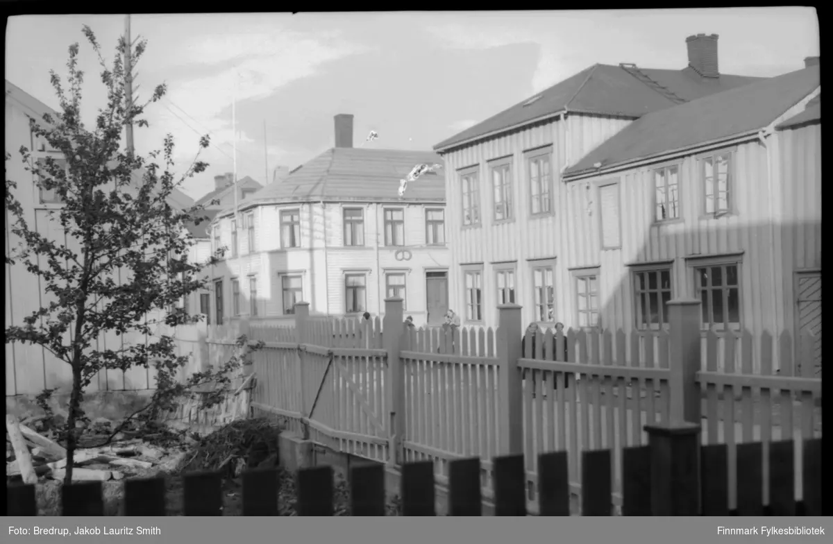 Fotografert i Vadsø sentrum. Vi ser bygninger, gater, en inngjerdet hage. I det første huset til høyre henger et bakeriskilt, dette er Henriksens bakeri i Hvistendahlsgate i Vadsø rundt 1920-tallet. Neste hus er Vinjevold gården. (Samme motiv som FBib.01009-093)