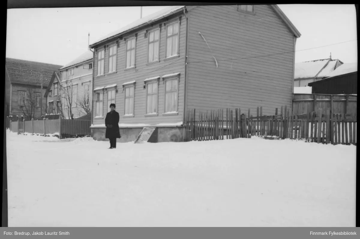 En mann står foran et toetasjers bolighus, antakelig i Inspektør Hansens gate i Vadsø.  Det er vinter, og hagene og gata er dekket av snø.  I bakgrunnen ser vi en del av Vadsø kirke.
