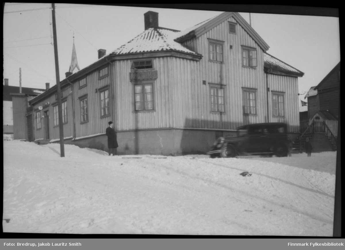 Bildet viser apoteket i Tollbugata, Vadsø.  Apoteket lå over Kildahl Olsens bokhandel.  En mann og en bil utenfor bygningen.
