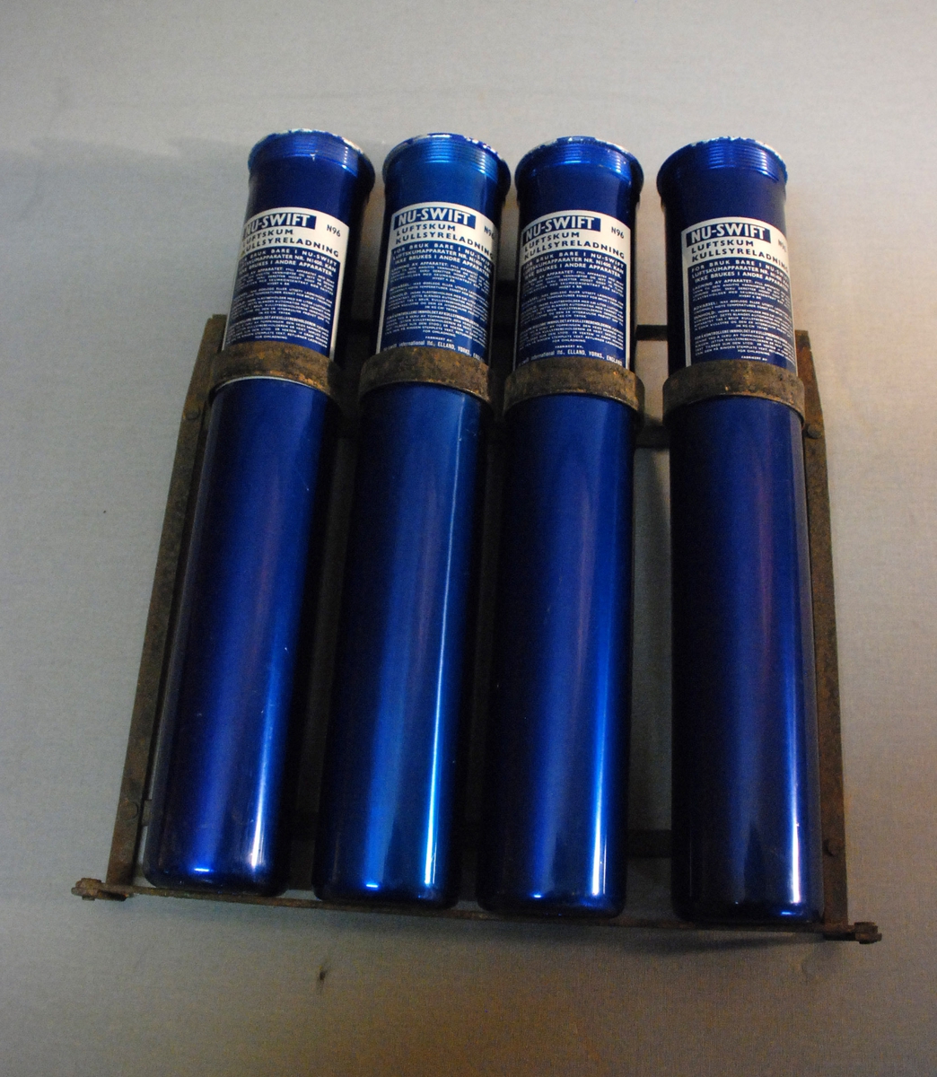 Metallstativ med fire  sylindriske patroner. Blå med hvit påtrykt tekst: Nu-swift Luftskum kullsyreladning M 96