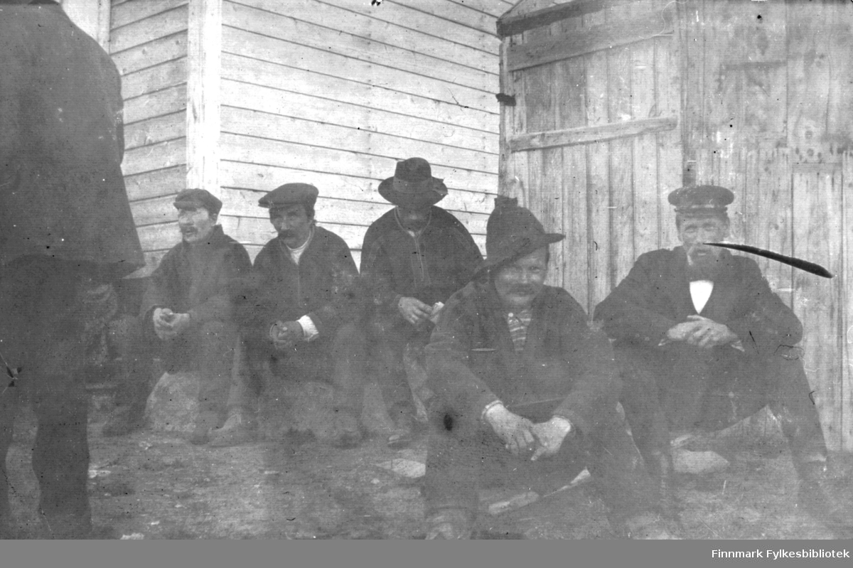 En gruppe menn sitter ved en husvegg, antakelig på Langnes, og kanskje i forbindelse med Langnesmarkedet. De fleste er koftekledde