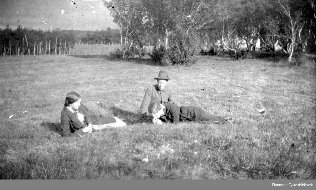 To damer og en mann ligger eller sitter på et jorde en fin augustdag. Mannen er Hans Gabrielsen. Mellom trærne i bakgrunnen kan man skimte et hus.