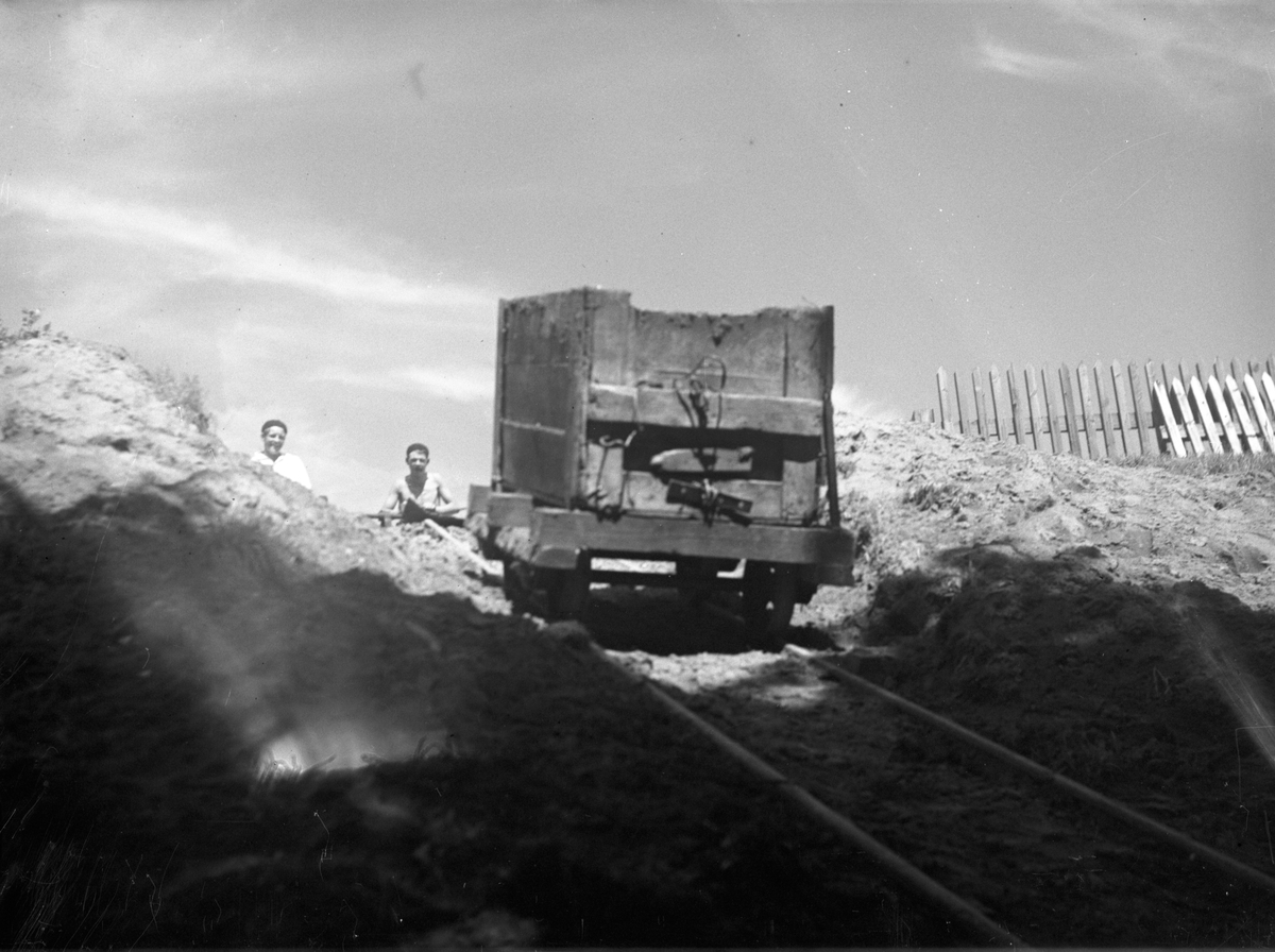 En vagn med en stor låda på rullar nerför backen på järnvägsspåretspåret, två pojkar tittar fram bakom sandkullarna.