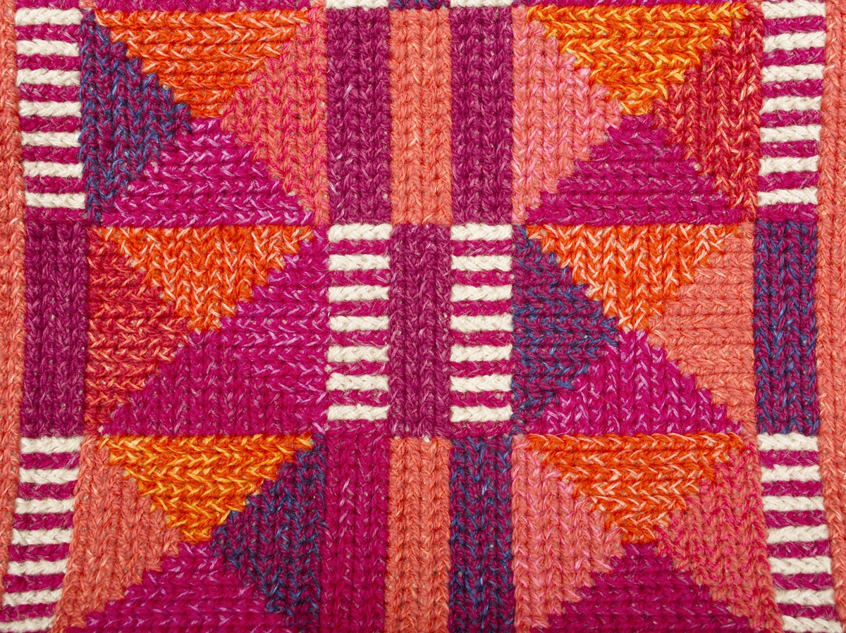 Handväska i tvistsöm med lingarn. Mönster av trianglar i olika röda och lila färger samt vitt. Lika fram- och baksida. Monterad med trähandtag, rött linnefoder. Formgivning av textilkonstnär Kerstin Mauritzson.
