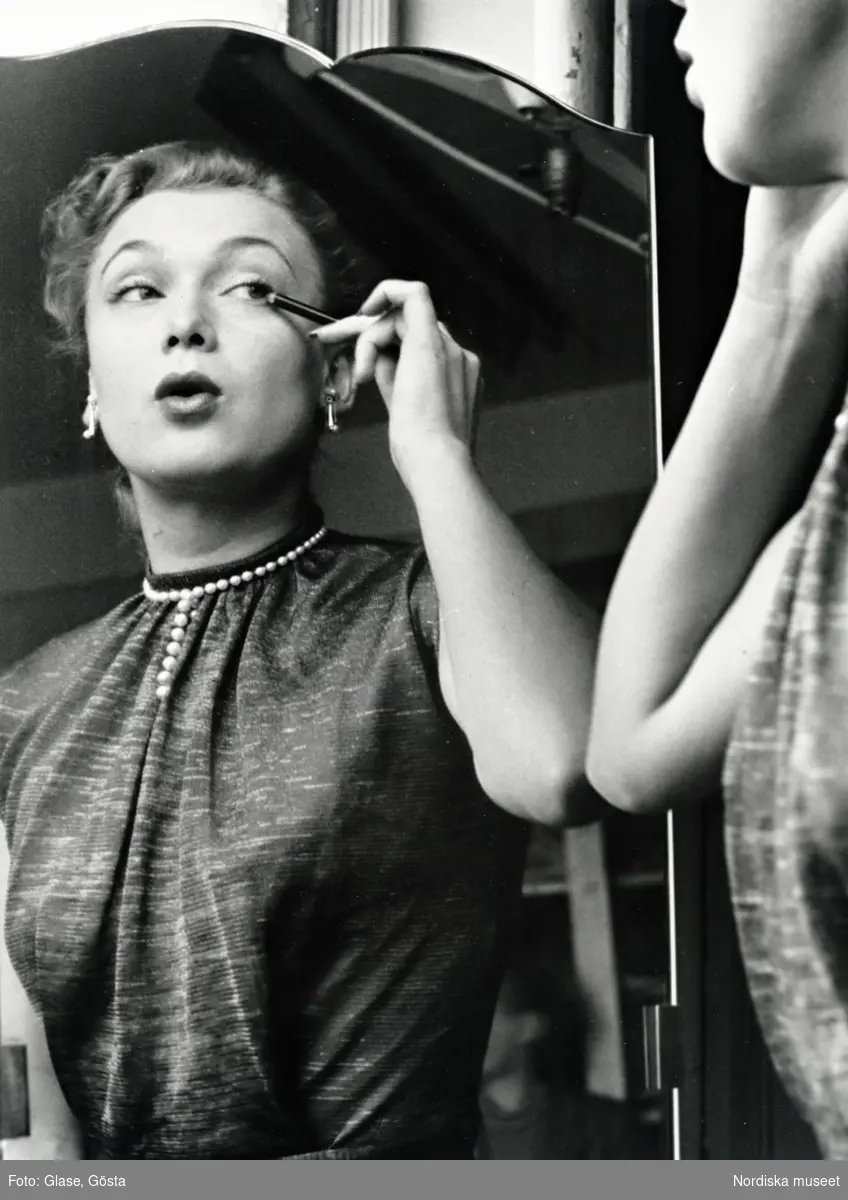 Dammode. Mannekängen Anita Sandin sminkar sig med kajalpenna och speglar sig i spegel. Örhängen och pärlhalsband.