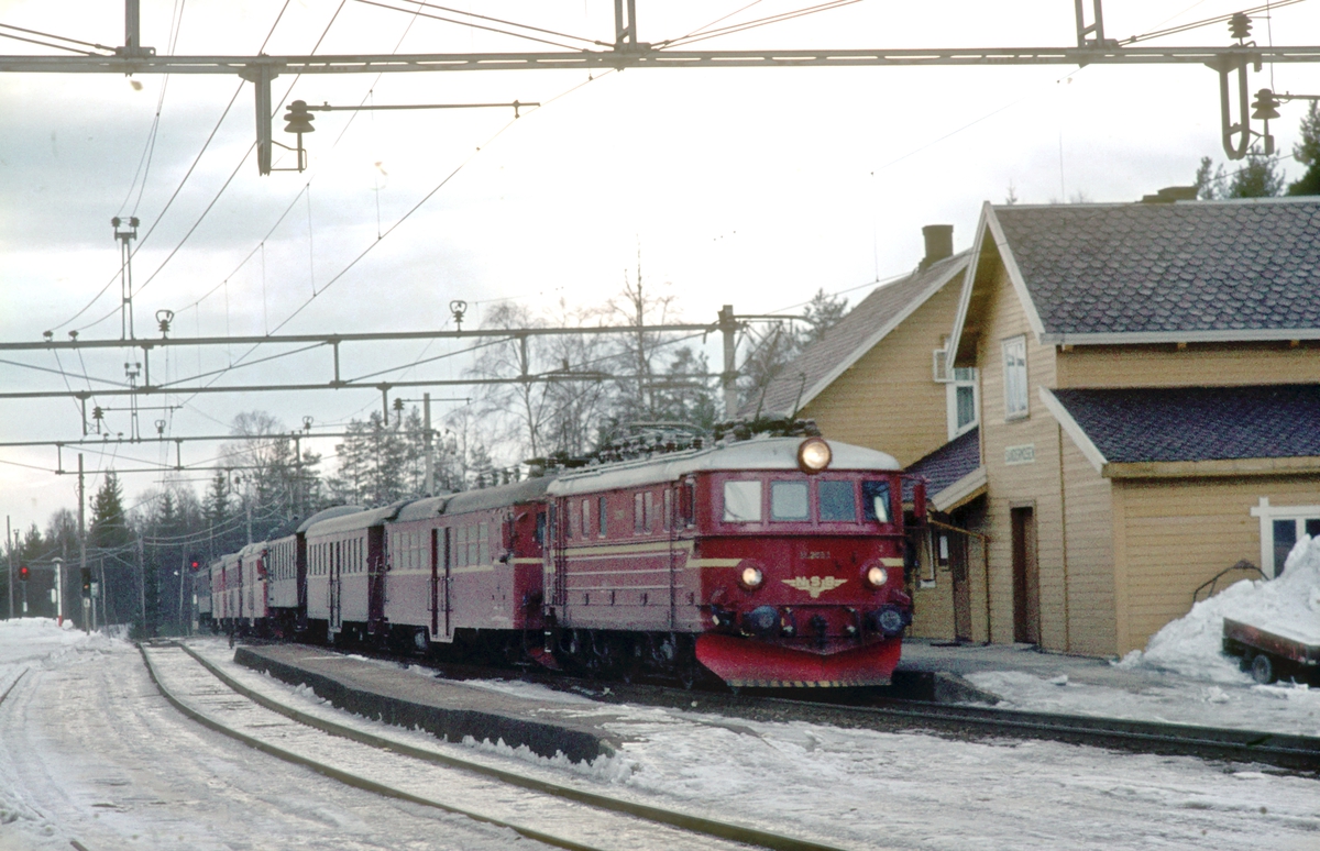 Persontog 207 Oslo Ø - Gjøvik passerer Sandermosen stasjon. NSB elektrisk lokomotiv type El 11 og to motorvognsett type 65, samt en enkel personvogn bakerst. På grunn av påsken var det mangel på personvogner og det gikk derfor motorvognsett (lokaltogsmateriell) i dette toget.