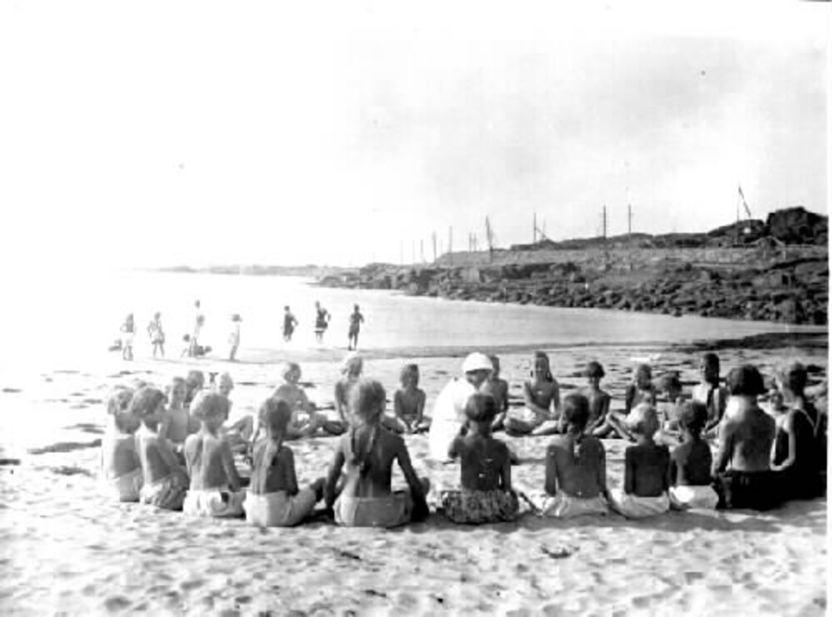 Barn från Kustsanatoriet Apelviken som badar och sitter i ring på stranden kring en ur personalen. Patienterna där led av körteltuberkulos.