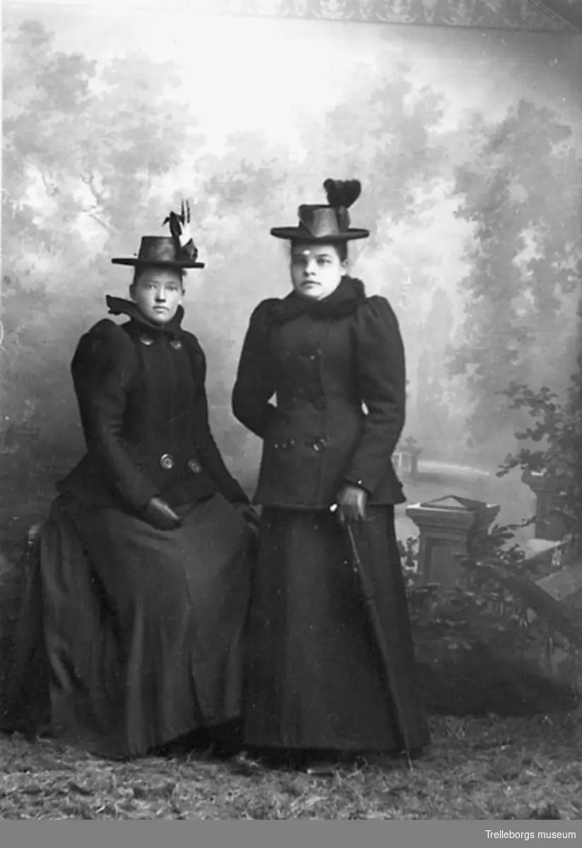 Två kvinnor i tvådelad dräkt med dubbelknäppt jacka, åtsittande ärmar med en lättare puff högst upp, handskar och högkullig hatt prydd med band och plymer. En med paraply vid sin sida.