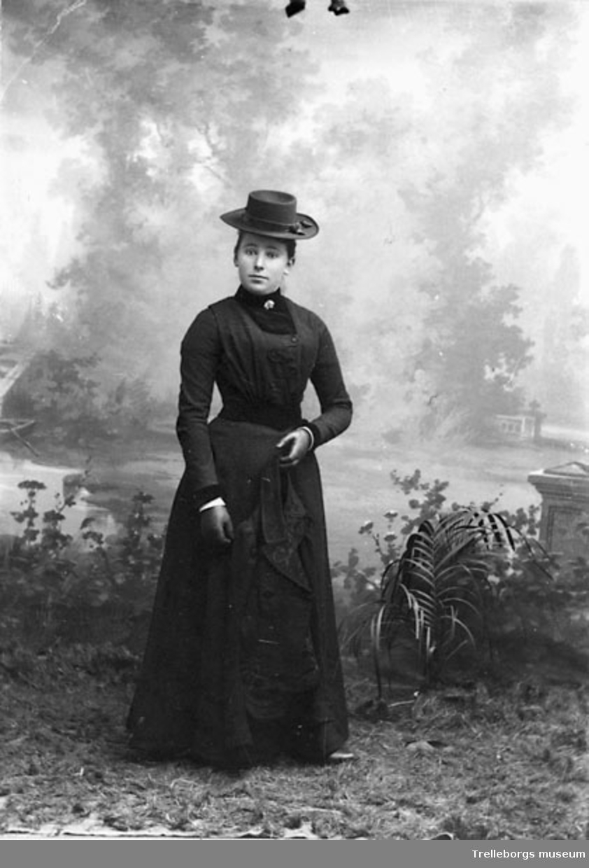 Kvinna i svart klänning  med broderad jacka, svarta handskar och hatt.