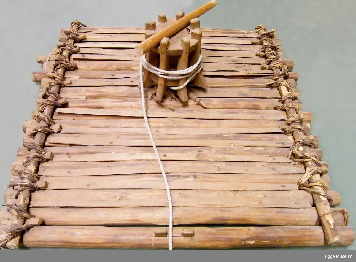 Modell av spillflåte laget i rundbarket gran. Flåten er bygd av rundtømmer som er kledd med halvklovninger. Tømmerstokkene er festet med vidjespenninger, som er kilt fast til stokker med store kiler stukket inn i løkker laget av vidjespenning. Ubehandlet. Flåten er tilnærmet kvadratisk i form. Midt på flåten står "jomfrua" eller tønna med oppkveilet tau. Det følger med en dreg laget av ståltråd. Ingen årer. Sammenføyningen med vidjebånd gjorde at flåten tålte bølger. Det er ukjent hvem som har laget modellen. Ukjent modellformat.