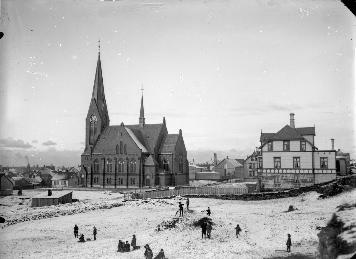 Vår Frelsers kirke en vinterdag. Flere barn i forgrunnen. En kjelke. Trehus til høyre, og trehusbebyggelse til venstre for kirken.