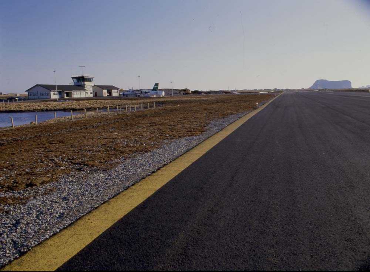 Lufthavn/Flyplass. Røst. Et fly, DHC-6 Twin Otter  fra Widerøe står parkert ved Ekspedisjonen/Flytårnet. I bakgrunnen ses Vedøya.