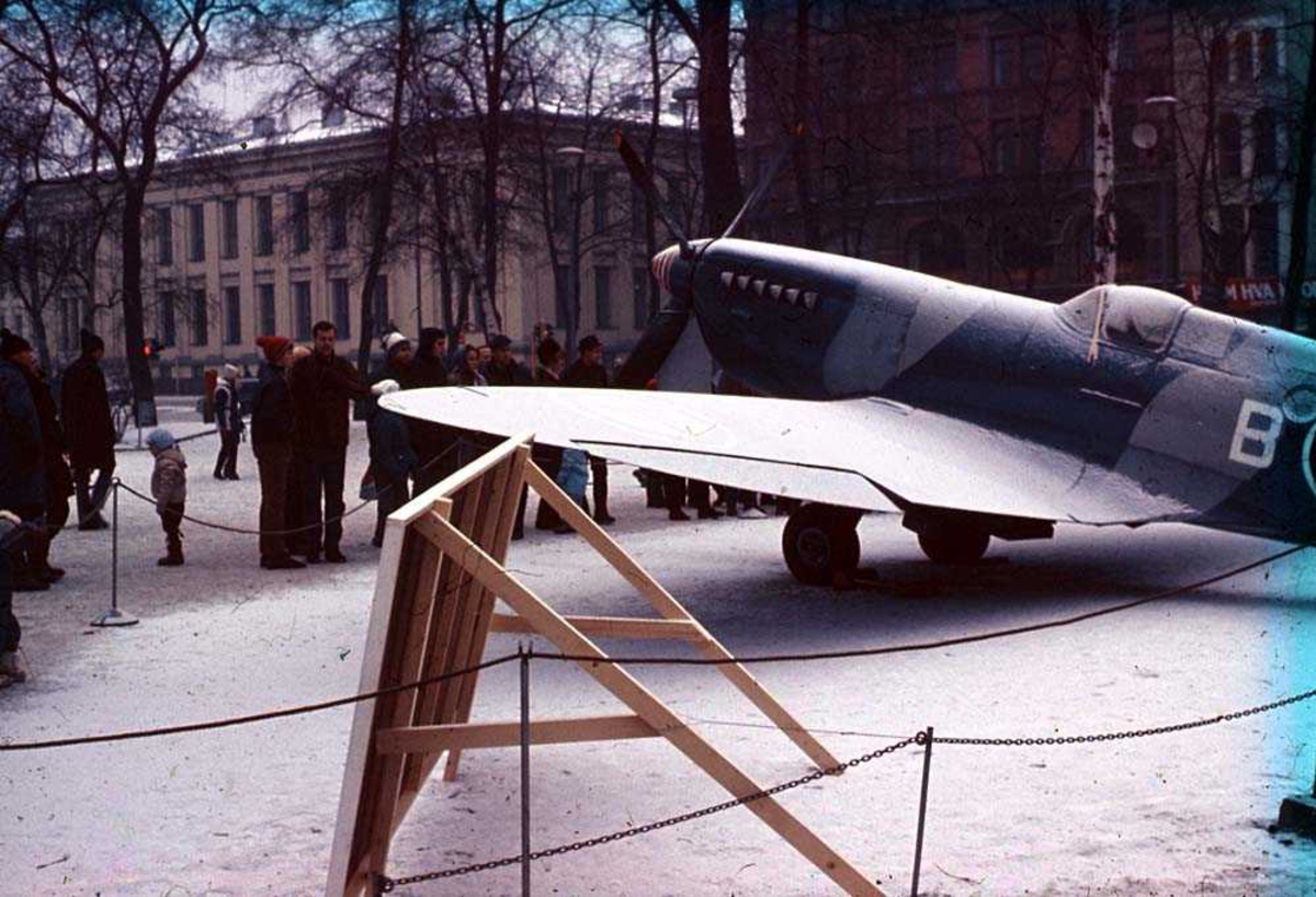 Ett fly som står utstillt i en park, Spirtfire Mk P. R. XI AZ-B. Flere personer og bygninger i bakgrunnen. Snø på bakken.