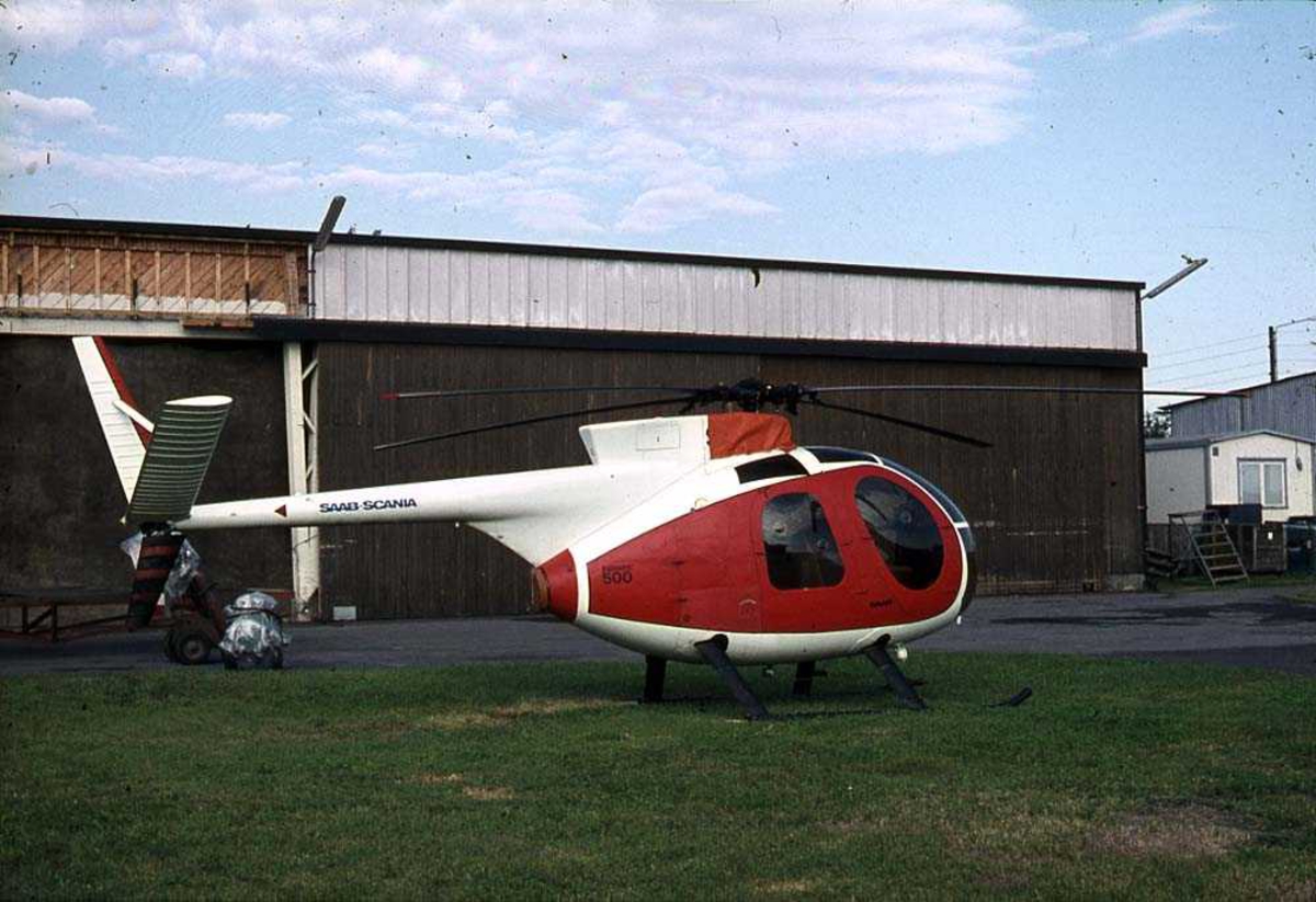 Ett helikopter på bakken, Hughes 500. Bygning i bakgrunnen.