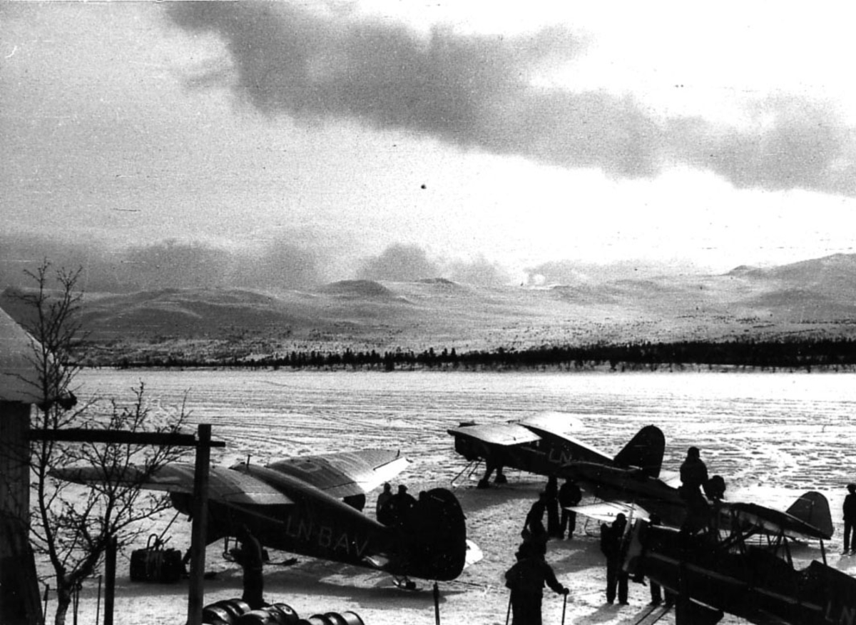 Åpen plass, tre fly på bakken med skiunderstell fra Widerøe. Flere personer ved flyene.