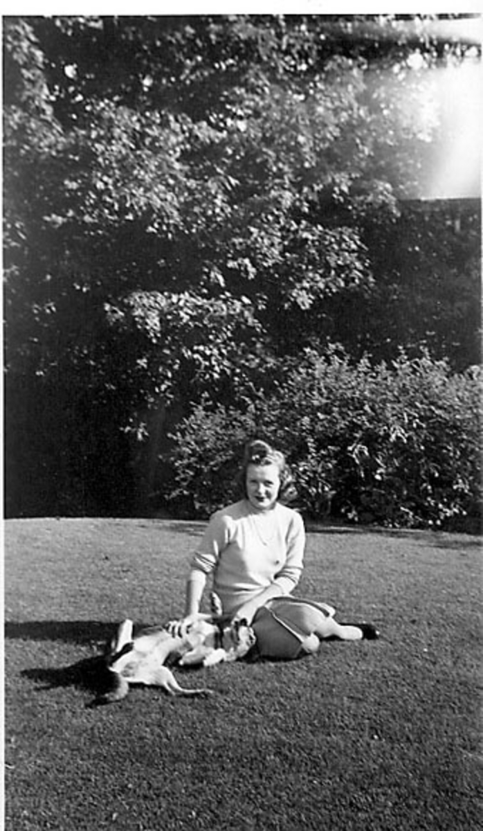 Portrett, person sitter på bakken, ant. i en park, og leker med en hund. Ung kvinne i sivile klær.