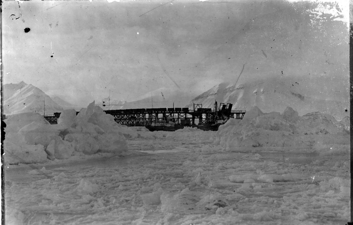 Kai med flere jernbanevogner. Fartøyet "Hobby" ligger ved kaia. Ismasser i forgrunnen, snø.