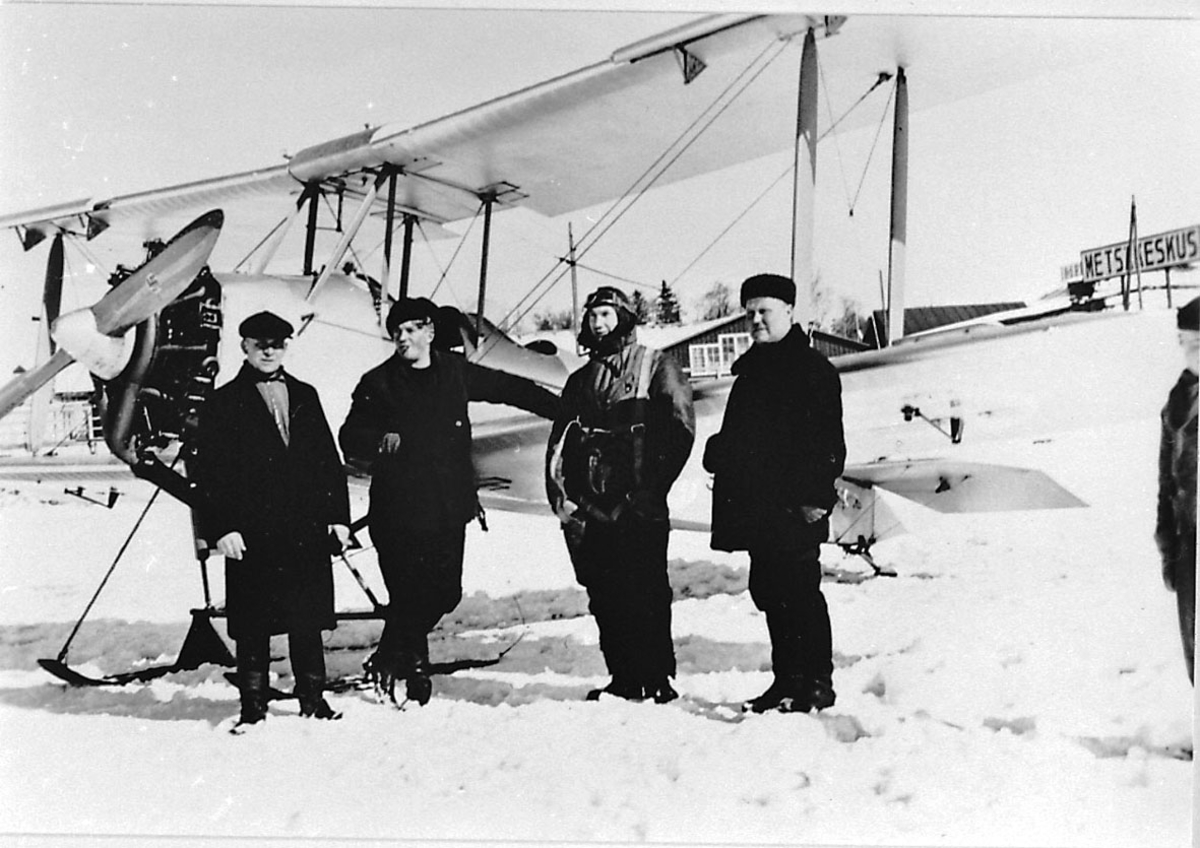 Fly, propellfly, dobbeldekker, Sääski II, 4 personer foran, 3 sivilkledde og en i flygerutstyr. Flyet med skiunderstell, snø på bakken. Bygninger bak.