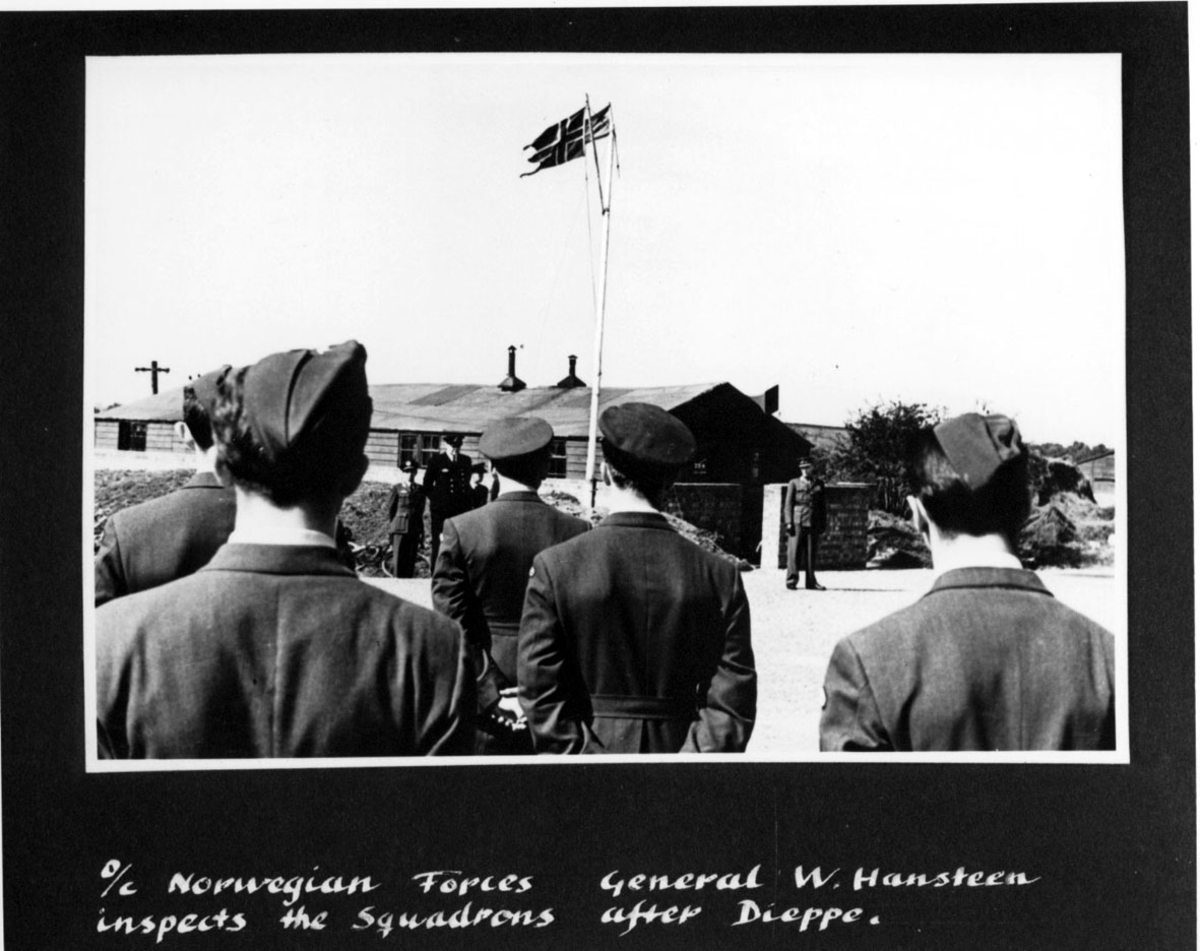 Flere personer i militæruniform, offiserer, soldater. Personene foran med ryggen i mot. Bak sees stang - mast med 2 flagg, og bygning. Håndskrevet engelsk tekst under bildet.