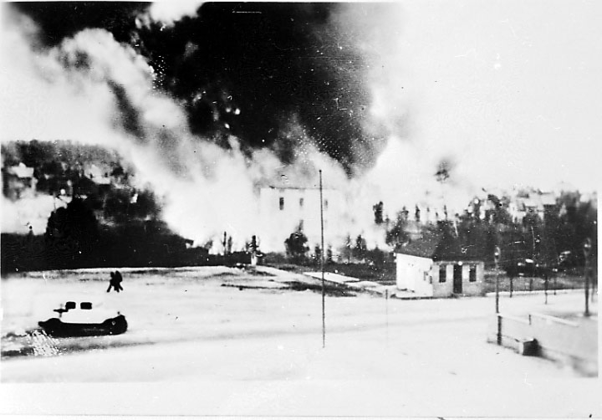 Røyk fra bygninger, i forgrunnen åpen plas med 2 personer. Narvik etter bombing - kriksødeleggelser, under 2. verdenskrig.