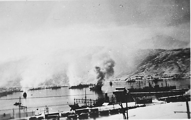 Havneområdet, flere fartøy, noen av dem står i brann, røyksøyler. Narvik under bombing, kriksødeleggelser, 2 verdenskrig.