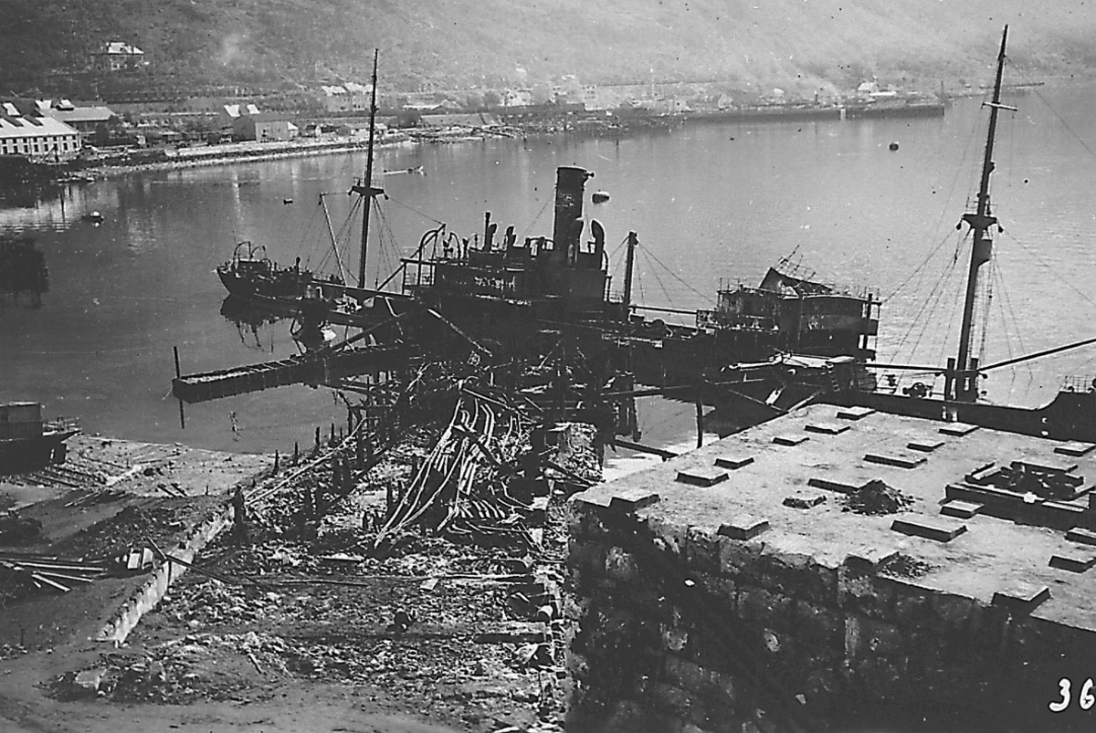 Tettsted/by. Havneområde med 2 fartøy. Havneområdet og fartøyene ødelagt etter bombeangrep. Bygninger i bakgrunnen.