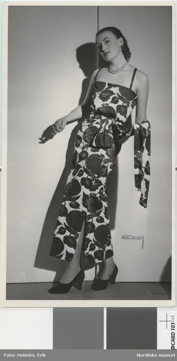 Nordiska Kompaniet. Kvinna i aftonklänning med tvär ringning, smala axelband och slits i sidan. Stort mönster med stiliserade granatäpplen och blad. Text på baksidan: "Molyneux uppvisning våren 1947."