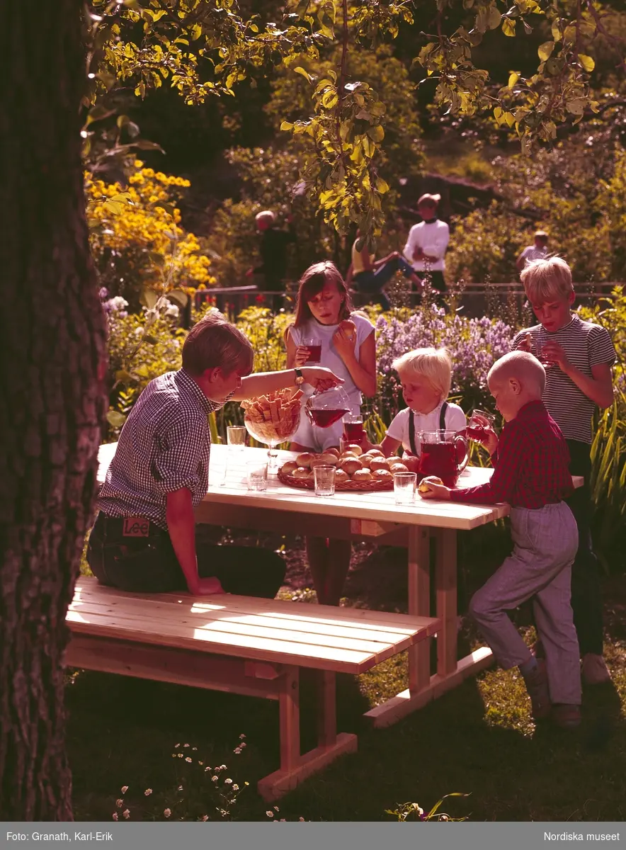 Ett sällskap av barn  runt ett dukat bord under ett träd. En av pojkarna häller upp saft. på bordet ett fat med bullar i bakgrunden lek.