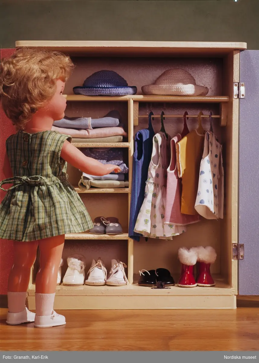 En docka som bläddrar i sin garderob med kläder , skor och hattar
Ur bildserien  "Dockans nya kläder" för tidningen Vi.