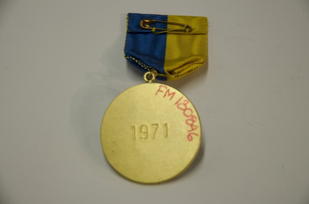 Idrottsmedalj i metall med stavar och skidor som motiv. Medaljen har ett blå, gult band och är från Tornedalsloppet. Den är färglad i ljusblått ovanpå gulmetallen. På baksidan är ingraverat 1971.