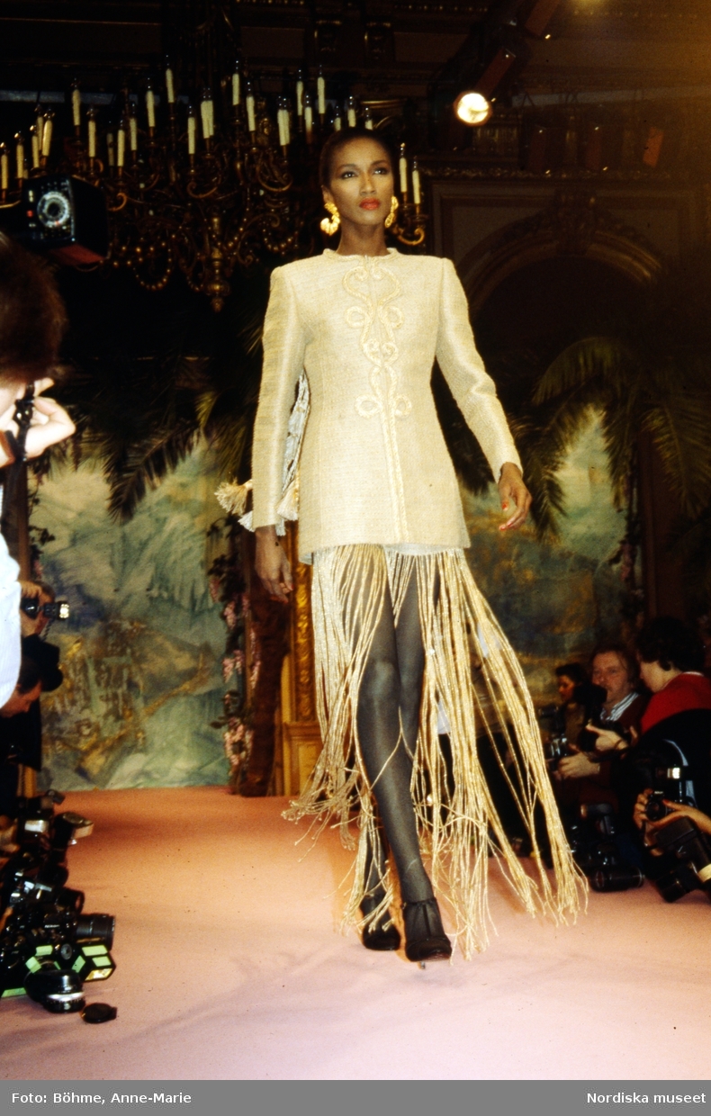 Modevisning. Modell i kort, guldfärgad klänning med fransar nedtill och örhängen. Från Lacroix.
